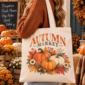 Könnte beinhalten: Eine cremefarbene Stofftasche mit einem Grafikdruck aus orangefarbenen und weißen Kürbissen, Herbstblättern, Beeren und Eicheln. Der Text "AUTUMN MARKET" ist in einer fetten, geschwungenen Schrift über der Ernteillustration gedruckt.
