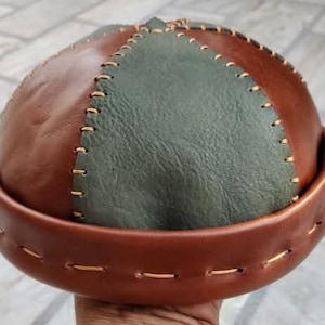 May include: A dome-shaped leather cap featuring alternating panels of smooth brown leather and textured olive green leather. The panels are joined by visible light-coloured stitching, and the cap includes a wide, folded brown leather brim.