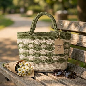 May include: A crochet tote bag featuring a two-tone olive green and cream wave pattern with a solid cream base and two handles. A brown paper tag attached to the handle displays the text Handmade With Love. A bouquet of daisies and sunglasses are nearby.