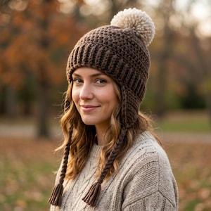 May include: A dark brown crocheted beanie featuring a ribbed brim, ear flaps, and two long braided tassels hanging from the sides. The hat is topped with a large, fluffy, cream-colored pom-pom made of yarn, providing a textured and warm appearance.