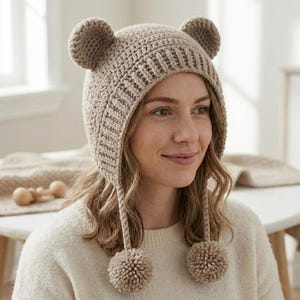 Puede incluir: Un gorro de ganchillo beige con dos orejas redondeadas similares a las de un oso en la parte superior y dos largas borlas trenzadas que cuelgan de los lados, cada una terminada con un gran pompón de hilo a juego. Tiene una textura acanalada.