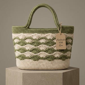 May include: A crochet tote bag featuring a two-tone olive green and cream color scheme with a wavy shell stitch pattern. The bag has two sturdy handles and a brown paper tag attached with string that reads, "Handmade With Love" in black script.