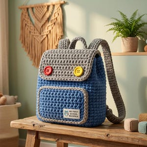 May include: A blue and gray crocheted backpack featuring a top flap secured by one red and one yellow button. The front pocket displays a white rectangular label with an owl graphic and the text MY FIRST SCHOOL BACKPACK in black lettering.