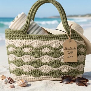 May include: A crochet tote bag featuring alternating olive green and cream wavy shell patterns. The bag contains a striped towel and a straw hat. A brown paper tag attached to the handle displays the text Handmade With Love in black script.