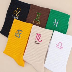 May include: A collection of six ribbed crew socks in black, brown, green, yellow, beige, and white. Each sock features a unique embroidered zodiac symbol in a contrasting thread colour, with some including additional embroidered text below the icon.