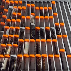 Peut inclure: Un organisateur de tiroir en métal gris comportant de nombreux compartiments verticaux séparés par des séparateurs gris et des étiquettes en plastique orange vif. Les compartiments sont remplis de forets métalliques de tailles variées, certains rangés dans de petits étuis en plastique transparent.