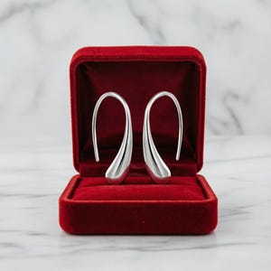 May include: A pair of polished silver-tone teardrop earrings with a curved hook design, presented inside a rectangular red velvet jewellery box with a hinged lid. The earrings feature a smooth, reflective metallic finish and a minimalist shape.
