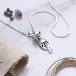 May include: A silver-toned necklace featuring a pendant shaped like a cat in a stretched, climbing pose. The pendant is attached to a delicate box chain. The cat figure has a smooth, reflective surface and a curved tail, creating a sleek appearance.