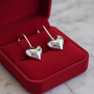 May include: A pair of polished silver-toned heart-shaped drop earrings with curved hooks, presented inside a rectangular red velvet jewellery box with a hinged lid. The earrings feature a smooth, reflective metallic surface and a rounded heart design.