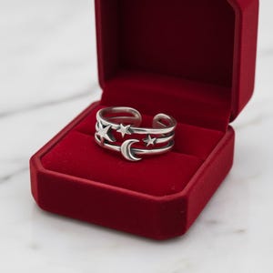 May include: A silver-toned open band ring featuring a celestial motif with a crescent moon and three stars. The ring is displayed inside a rectangular, deep red velvet-lined jewellery box with a hinged lid and a smooth, matte exterior finish.