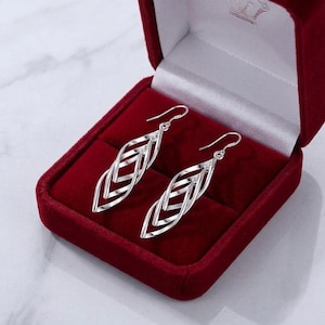 May include: A pair of silver-toned drop earrings featuring an elongated, multi-layered marquise shape with a twisted, open-work design. The earrings are suspended from curved ear wires and are displayed inside a deep red velvet jewellery box.