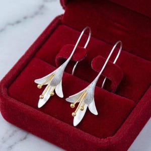 May include: A pair of drop earrings shaped like lily flowers, featuring textured silver petals and gold-toned stamens with small spherical tips. The earrings have long, curved silver ear wires and are presented inside a deep red velvet jewellery box.
