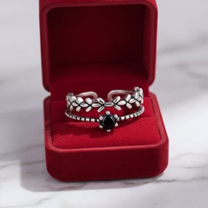 May include: A silver-toned open ring featuring two bands: one with a repeating leaf-like pattern and another with a central black circular stone set in a six-prong mount. The ring is displayed inside a square, bright red velvet jewellery box.