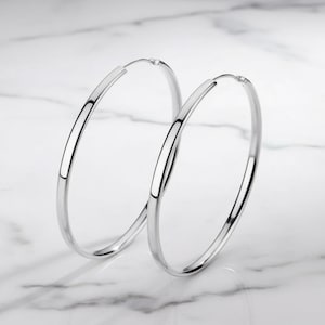May include: A pair of large, polished silver-tone hoop earrings. These circular earrings feature a smooth, reflective metallic finish and a thin, lightweight construction. The design is simple and classic, consisting of two continuous metal loops.
