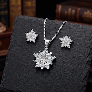 May include: A silver-toned jewellery set consisting of a pendant necklace and a pair of matching stud earrings. Each piece features a flower-shaped design composed of multiple clear, round-cut stones arranged around a central metallic point.