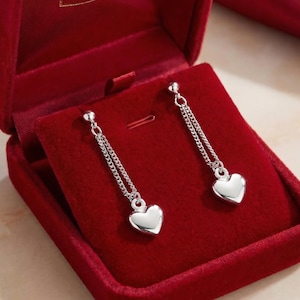 May include: A pair of silver-toned drop earrings featuring a delicate chain design that suspends a smooth, polished heart-shaped charm. The earrings are presented against a deep red velvet-lined jewellery box, highlighting the metallic finish.