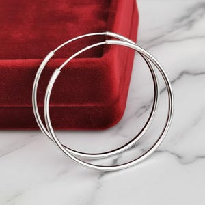 May include: A pair of large, polished silver hoop earrings with a thin, circular wire design. The earrings feature a simple, continuous loop construction with a smooth, reflective metallic finish, presented as a set of two identical circular hoops.