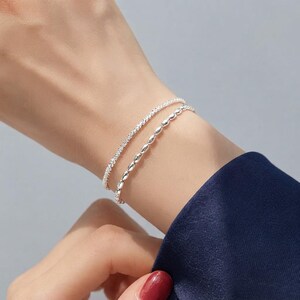 May include: A double-strand silver bracelet featuring one thin chain with a textured, sparkling finish and a second chain composed of small, smooth, oval-shaped metallic beads. The two delicate silver strands rest together on the wrist.