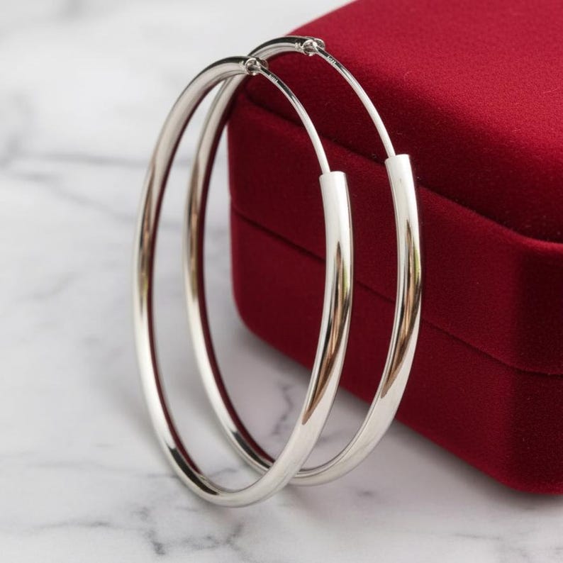 May include: A pair of large, polished silver hoop earrings with a smooth, rounded profile. The earrings feature a thin, continuous circular design with a simple wire closure mechanism, showcasing a bright, reflective metallic finish throughout.