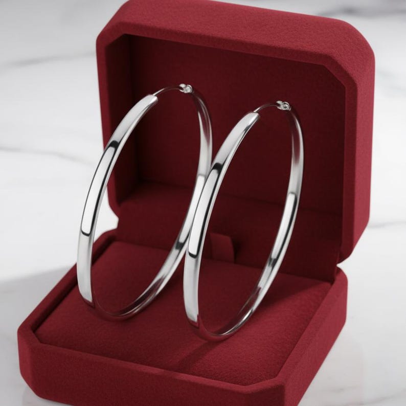 May include: A pair of polished silver hoop earrings with a smooth, rounded profile. The earrings are displayed upright inside an open, deep red velvet-lined jewelry box with a hinged lid, showcasing their reflective metallic surface and circular shape.