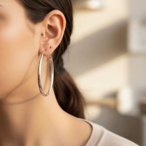 May include: A large, polished silver hoop earring worn in the earlobe, featuring a smooth, circular design. The earring is complemented by two small, simple silver stud earrings positioned higher up on the ear cartilage, creating a cohesive look.