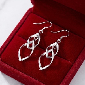 May include: A pair of silver-toned drop earrings featuring a polished, twisted metal design that forms an elongated, open oval shape. Each earring is suspended from a simple curved hook wire, showcasing a smooth, reflective surface throughout.