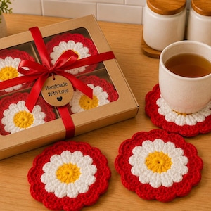 May include: A set of four round crochet coasters featuring a daisy flower pattern with a yellow center, white petals, and a red scalloped border. The coasters are packaged in a brown cardboard box with a clear window, tied with a red ribbon and a tag reading Handmade With Love.