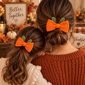 Könnte beinhalten: Zwei passende Häkel-Haarschleifen in Kürbisform mit leuchtend orangefarbenem Rippstoff, kleinen grünen Blättern und einem dunkelbraunen Stiel. Diese Haaraccessoires zeichnen sich durch eine strukturierte, handgefertigte Garnkonstruktion aus.