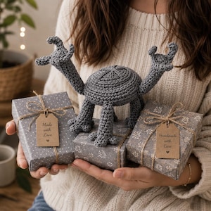 Puede incluir: Una criatura de ganchillo gris con cuatro patas y dos brazos levantados, colocada sobre tres cajas de regalo envueltas en papel gris estampado. Dos cajas tienen etiquetas de papel marrón con el texto Made with Love y Handmade with Care.