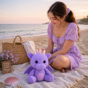 May include: A purple crocheted dragon plush toy with large black eyes, small wings, and a striped belly. The dragon features light-coloured horns and pink blush accents on its cheeks. It is crafted with a textured yarn finish and a rounded shape.