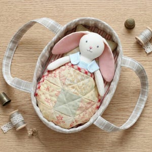 May include: A plush rabbit toy with long pink ears and a light blue top, tucked into a fabric-lined oval basket with handles. The rabbit is covered by a small, quilted patchwork blanket featuring floral patterns, alongside two thimbles and two spools.