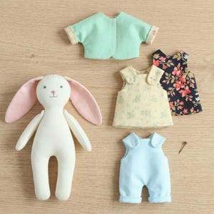May include: A cream-colored plush rabbit toy with long pink-lined ears, accompanied by a set of doll clothing including a mint green short-sleeve top, a yellow floral sleeveless top, a dark blue floral sleeveless top, and light blue overalls.