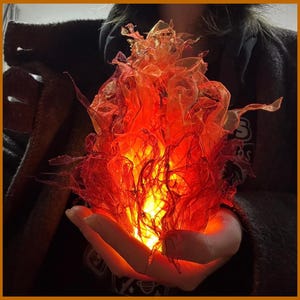 May include: A sculptural flame-shaped light fixture crafted from translucent, layered material in vibrant shades of red, orange, and yellow. The structure emits a warm, internal glow, mimicking the appearance of a flickering fire with jagged edges.