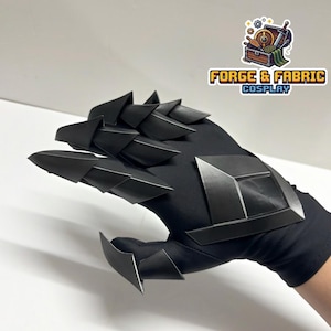 Puede incluir: Un guante negro con placas de armadura articuladas, superpuestas y puntiagudas que cubren los dedos y una placa geométrica en forma de escudo en el dorso de la mano. El logotipo de Forge & Fabric Cosplay es visible en la esquina superior derecha.