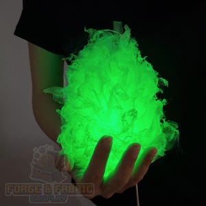May include: A glowing, bright green, textured, flame-shaped prop made of layered, translucent, ruffled fabric. The object emits a vibrant light from within, creating a sharp contrast against the dark background and the dark clothing worn.