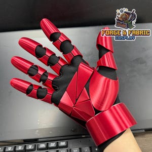Op de afbeelding: Een rode gearticuleerde handschoen met een zwarte stoffen basis en gesegmenteerde metallic rode platen die de vingers, handpalm en pols bedekken. De platen zijn in geometrische patronen gerangschikt. Het logo Forge & Fabric Cosplay is zichtbaar.