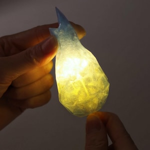 May include: A translucent, teardrop-shaped object with a faceted surface texture. The top section is a cool blue, while the lower, wider portion glows with a warm, bright yellow light emanating from within, creating a distinct color gradient.