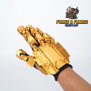 May include: A gold-colored articulated mechanical glove featuring segmented finger plates and a structured back plate with geometric detailing. The glove is constructed with a black fabric base and includes the text FORGE & FABRIC COSPLAY.