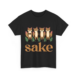 May include: A black short-sleeve t-shirt featuring a graphic of four brown foxes standing among green fern leaves. Below the foxes, the word "sake" is printed in large, bold, tan-colored letters. The design is centered on the front of the shirt.