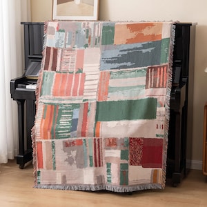 May include: A rectangular woven throw blanket featuring a multi-coloured abstract block pattern in shades of green, orange, red, blue, and cream. The textile has a textured appearance with frayed edges along the top and bottom borders.