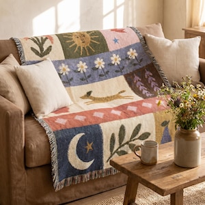 Puede incluir: Una manta tejida que presenta un diseño de varios paneles con ilustraciones de un sol, un perro corriendo, una luna creciente, flores y hojas en tonos azul, verde, crema y terracota, terminada con un borde de flecos en los laterales.