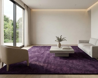 May include: A large, deep purple area rug with a textured, shaggy pile surface. The rectangular rug is positioned in a room featuring a light-coloured sofa, a beige armchair, and a low, square, light-grey coffee table topped with a small vase.