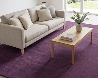 May include: A large, rectangular, deep purple area rug with a soft, plush texture. The rug is positioned on a light-coloured wooden floor, extending underneath a beige sofa and a light wood coffee table, providing a bold, solid colour foundation.