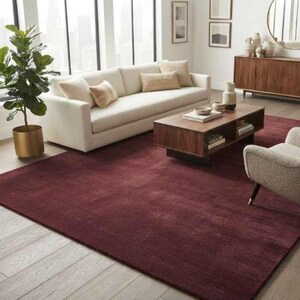 Burgundy wine red area rug styled in a modern living room featuring a cream boucle sofa, walnut wood coffee table, round gold mirror, and floor-to-ceiling windows with a city skyline view, showcasing the rug's rich color against light wood flooring.
