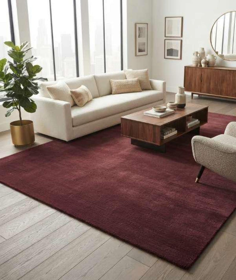 Burgundy wine red area rug styled in a modern living room featuring a cream boucle sofa, walnut wood coffee table, round gold mirror, and floor-to-ceiling windows with a city skyline view, showcasing the rug's rich color against light wood flooring.