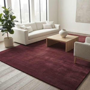Deep burgundy shag area rug in a minimalist living room with a cream sofa, light wood coffee table, ceramic vase, and floor-to-ceiling windows, emphasizing the rug's plush texture and rich wine red color against pale hardwood floors.