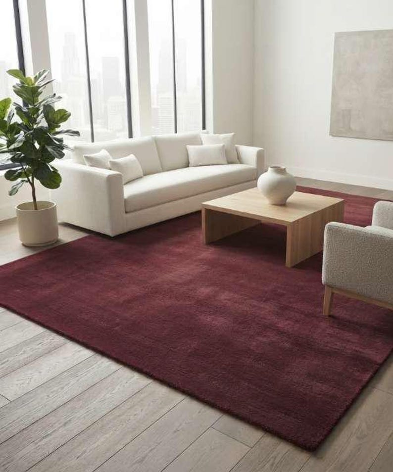 Deep burgundy shag area rug in a minimalist living room with a cream sofa, light wood coffee table, ceramic vase, and floor-to-ceiling windows, emphasizing the rug's plush texture and rich wine red color against pale hardwood floors.