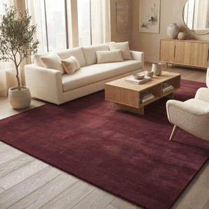 Wine red plush area rug in a warm neutral living room with a cream sofa, two boucle accent armchairs, light oak coffee table and console, potted olive tree, and soft natural lighting highlighting the rug's cozy texture and deep tone.