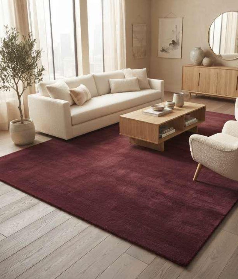Wine red plush area rug in a warm neutral living room with a cream sofa, two boucle accent armchairs, light oak coffee table and console, potted olive tree, and soft natural lighting highlighting the rug's cozy texture and deep tone.