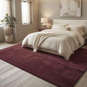 Burgundy wine red area rug in a cozy bedroom setting with a cream upholstered bed, knit throw blanket, walnut nightstands, and soft ambient lighting, highlighting the rug's soft plush pile and warm deep red tone underfoot.